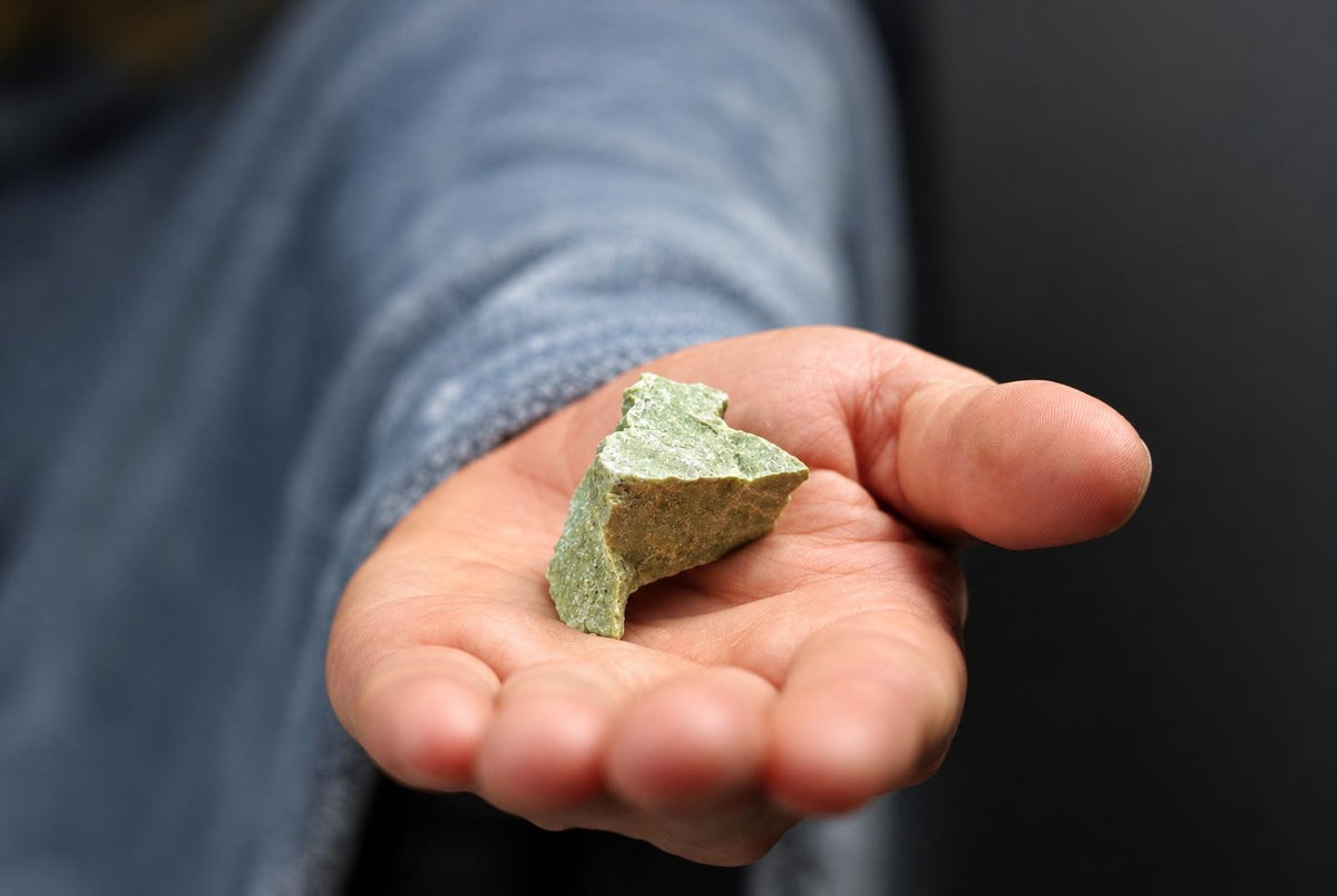 A hand holds a small, rough green stone on a dark surface.