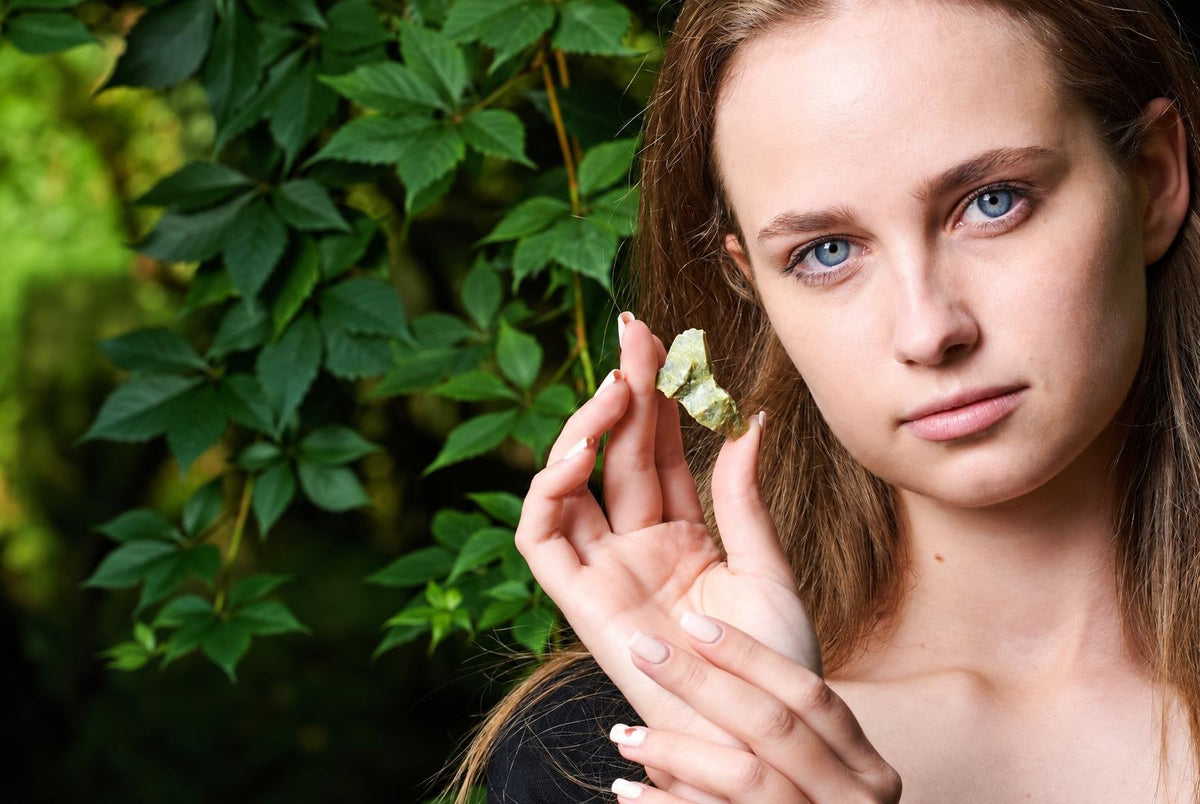 A woman holds a small, pale green raw stone against a dark surface with green leaves in the background.