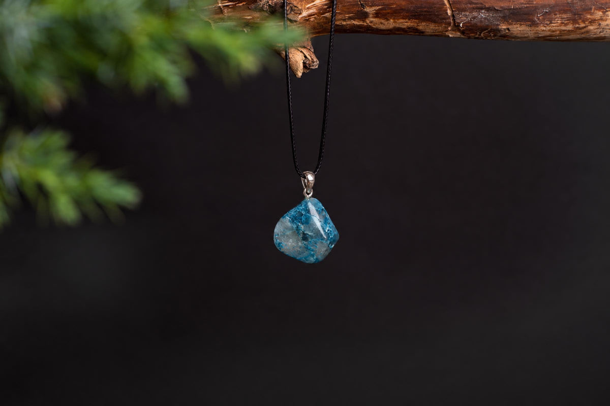 A blue and white speckled Shattuckite pendant hangs from a black cord on a dark surface, with green foliage in the background.