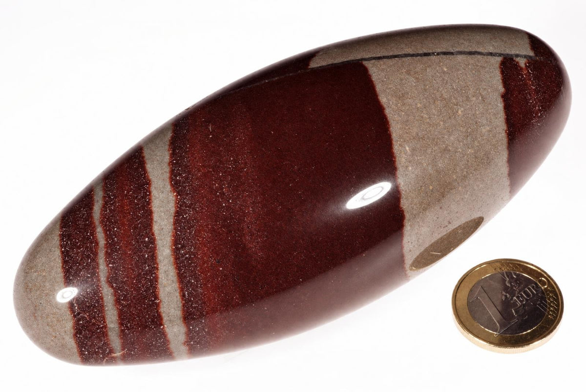 A polished, oval-shaped stone with dark red and grey stripes rests on a white surface next to a one-euro coin.