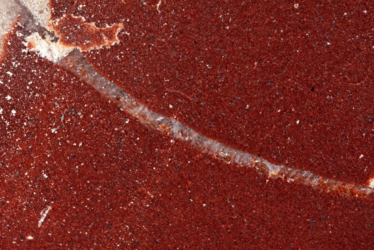 A close-up of a polished red stone surface with white speckles and a diagonal light-colored vein.
