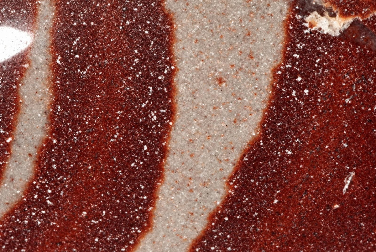 A polished stone surface with wavy bands of dark red and pale grey, speckled with white and black mineral grains.