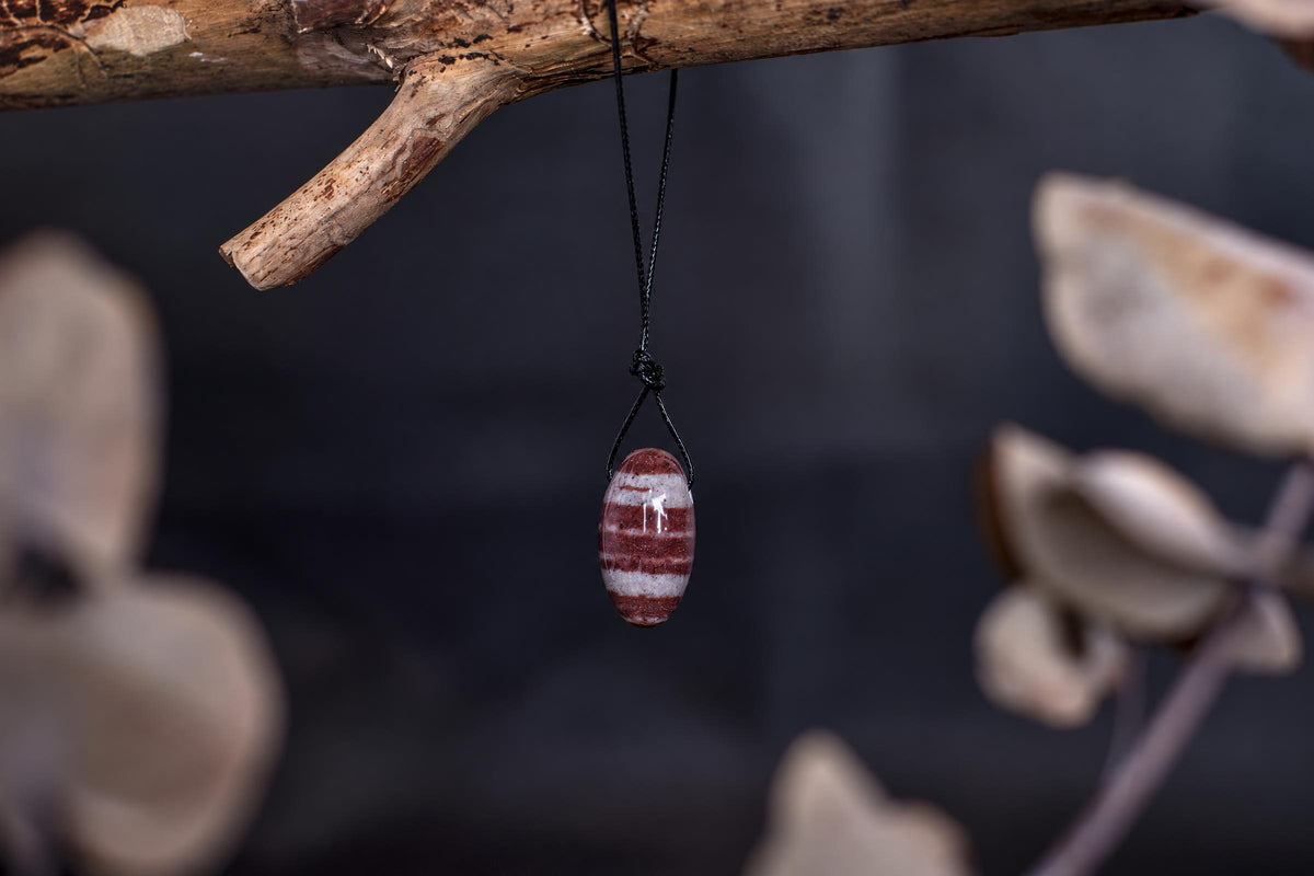A polished, oval-shaped pendant with horizontal red and white stripes hangs from a black cord on a wooden branch.