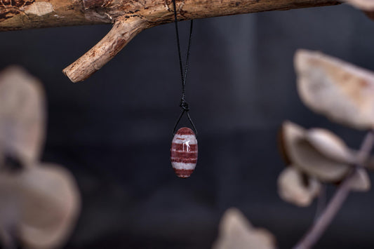 A polished, oval-shaped pendant with horizontal red and white stripes hangs from a black cord on a wooden branch.