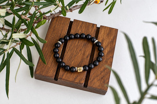 A bracelet of black round beads with a small square metal bead rests on wooden slats beneath narrow green leaves.