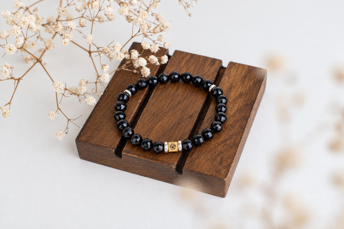 A bracelet with black faceted beads and a gold-toned square charm rests on a dark wooden stand beside dried flowers.