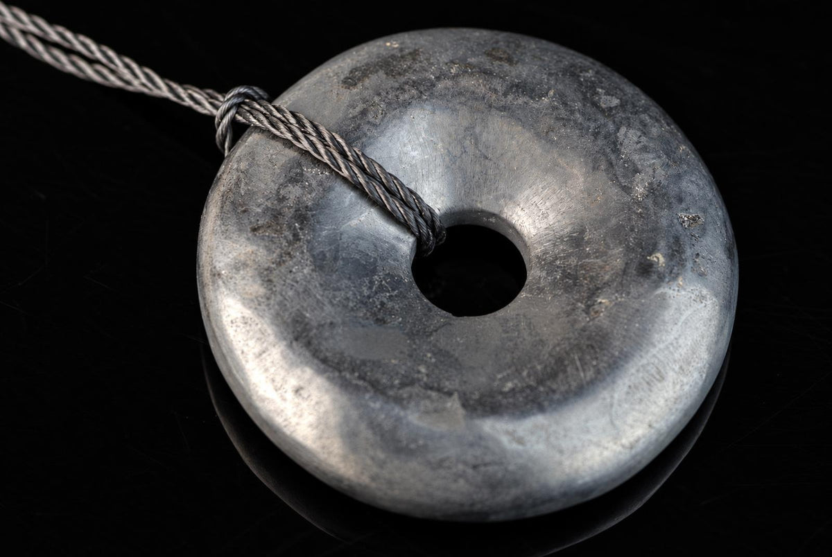 A silver-toned, donut-shaped pendant with a central hole and a braided cord looped through it, resting on a dark surface.