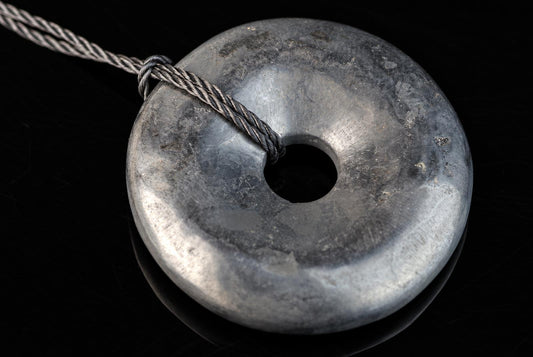 A silver-toned, donut-shaped pendant with a central hole and a braided cord looped through it, resting on a dark surface.