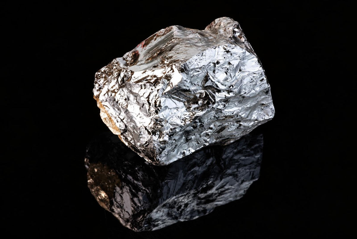 A rough, silver-toned block of silicon polycrystalline material rests on a dark, reflective surface against a black background.