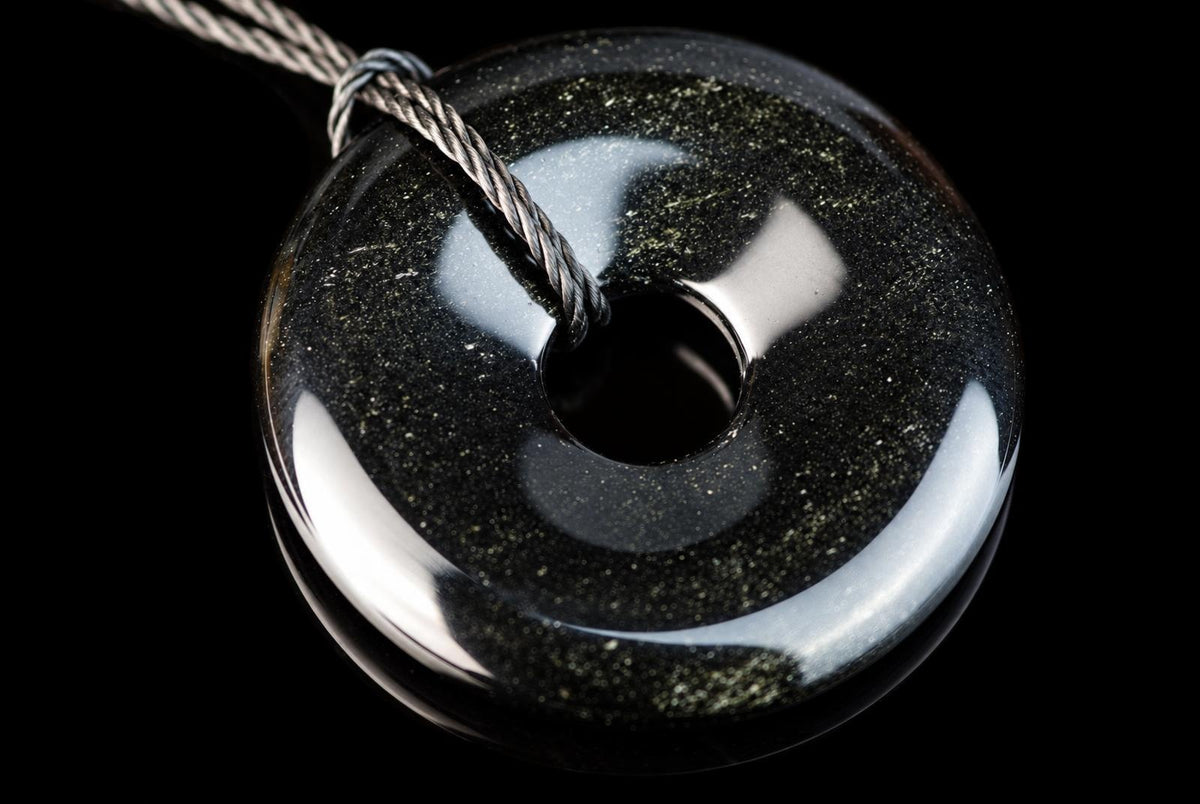 A polished black circular pendant with a central hole and fine pale speckles lies on a reflective black surface with a cord through its center.