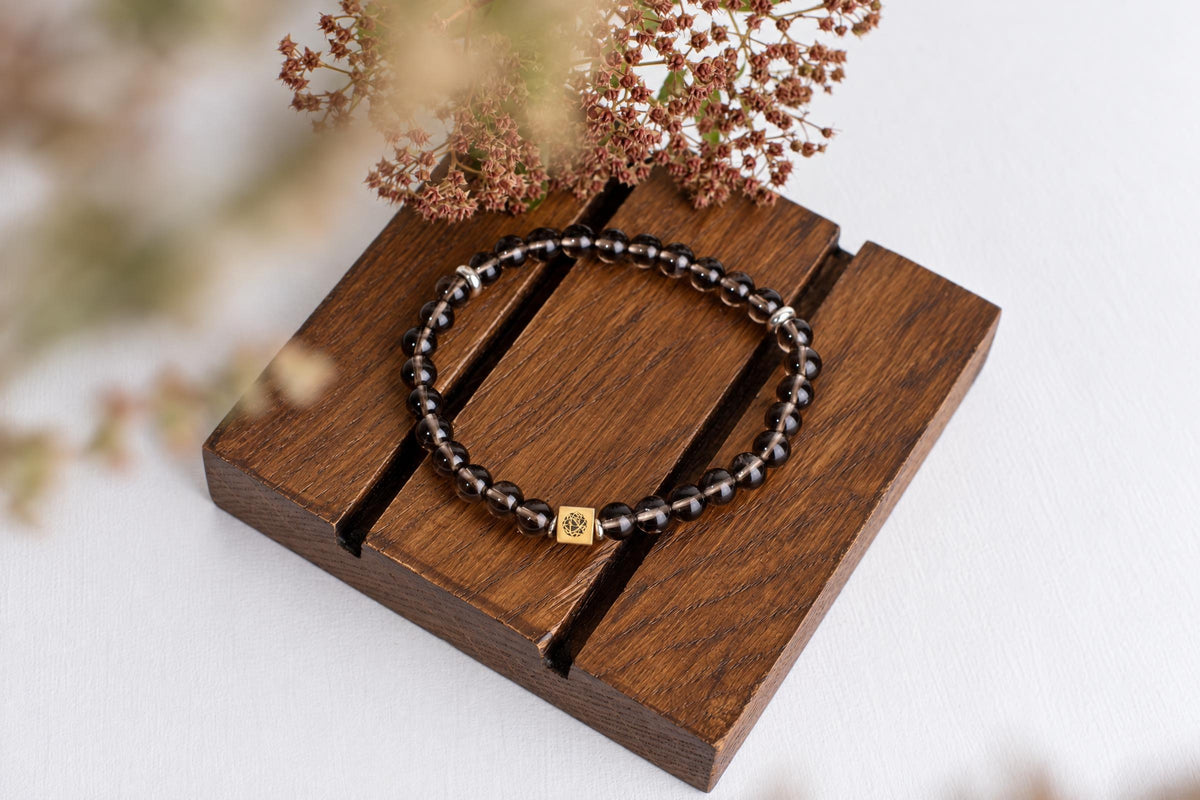 A bracelet of translucent dark brown round beads with a small square metal bead rests on wooden slats beside dried flower clusters.
