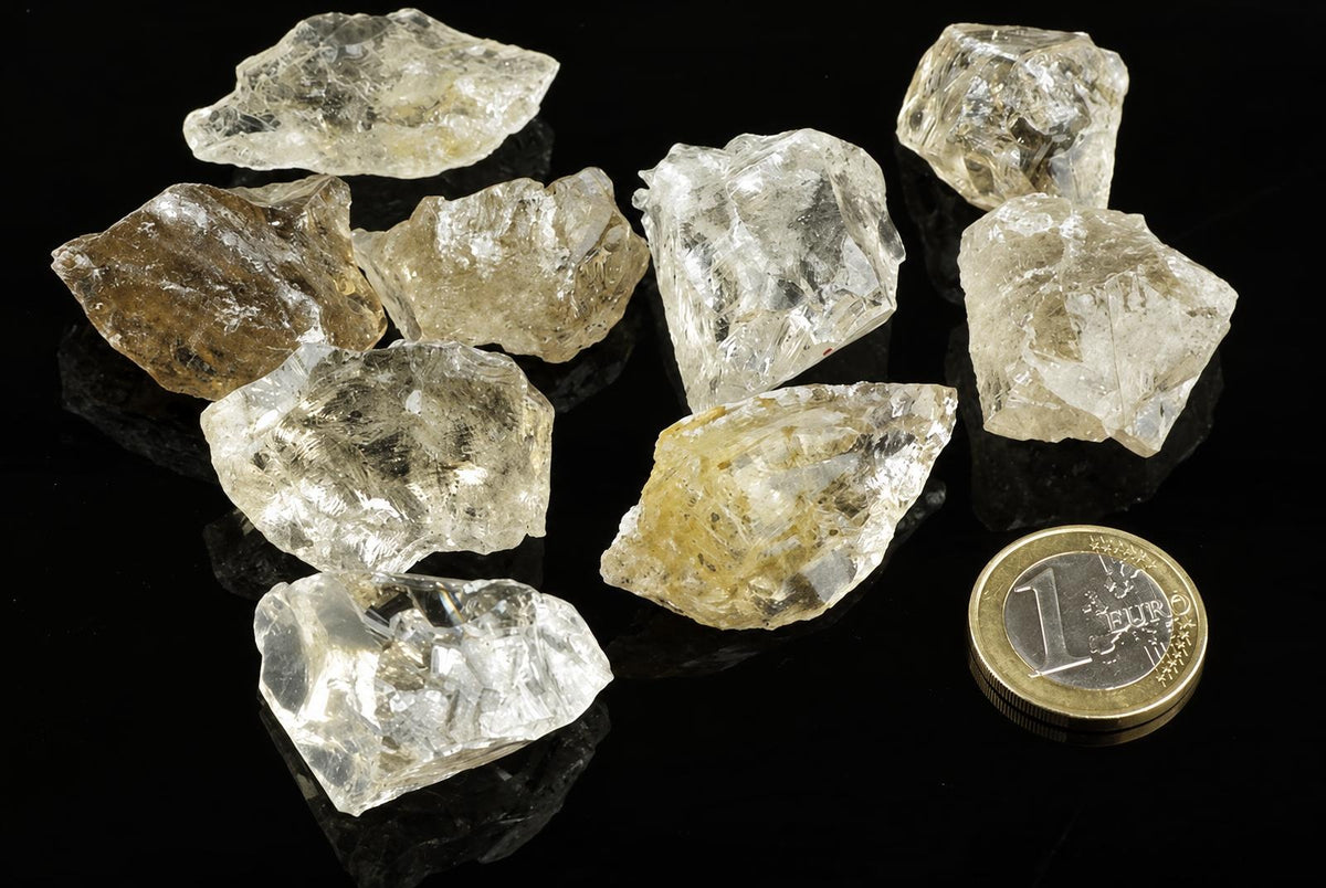 Several raw quartz crystals, mostly clear to pale yellow, on a dark surface with a one-euro coin for scale.