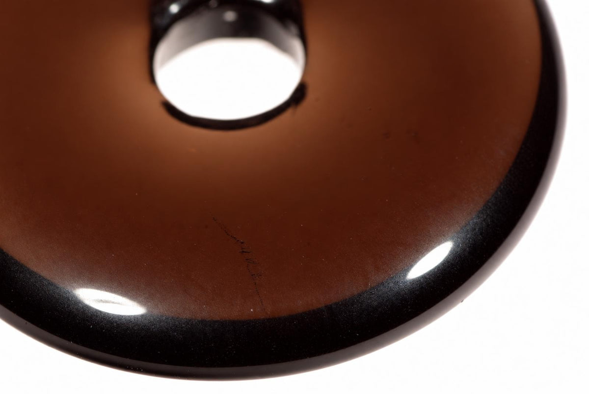 A close-up of a brown, donut-shaped pendant with a black rim and central hole on a white surface.