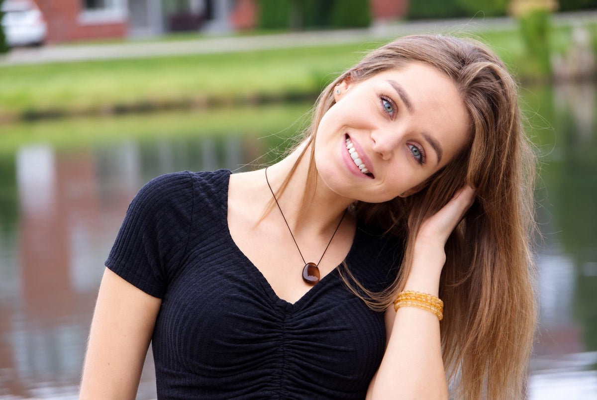 A smiling woman with long brown hair wears a black top, a dark pendant on a thin cord, and yellow bracelets near water.