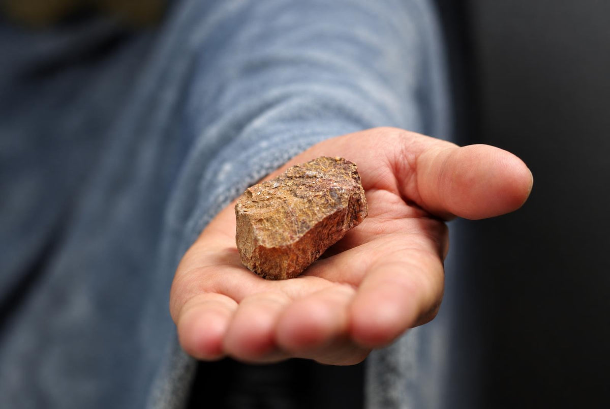 A hand holds a rough, brownish-red stone with a textured surface against a dark background.