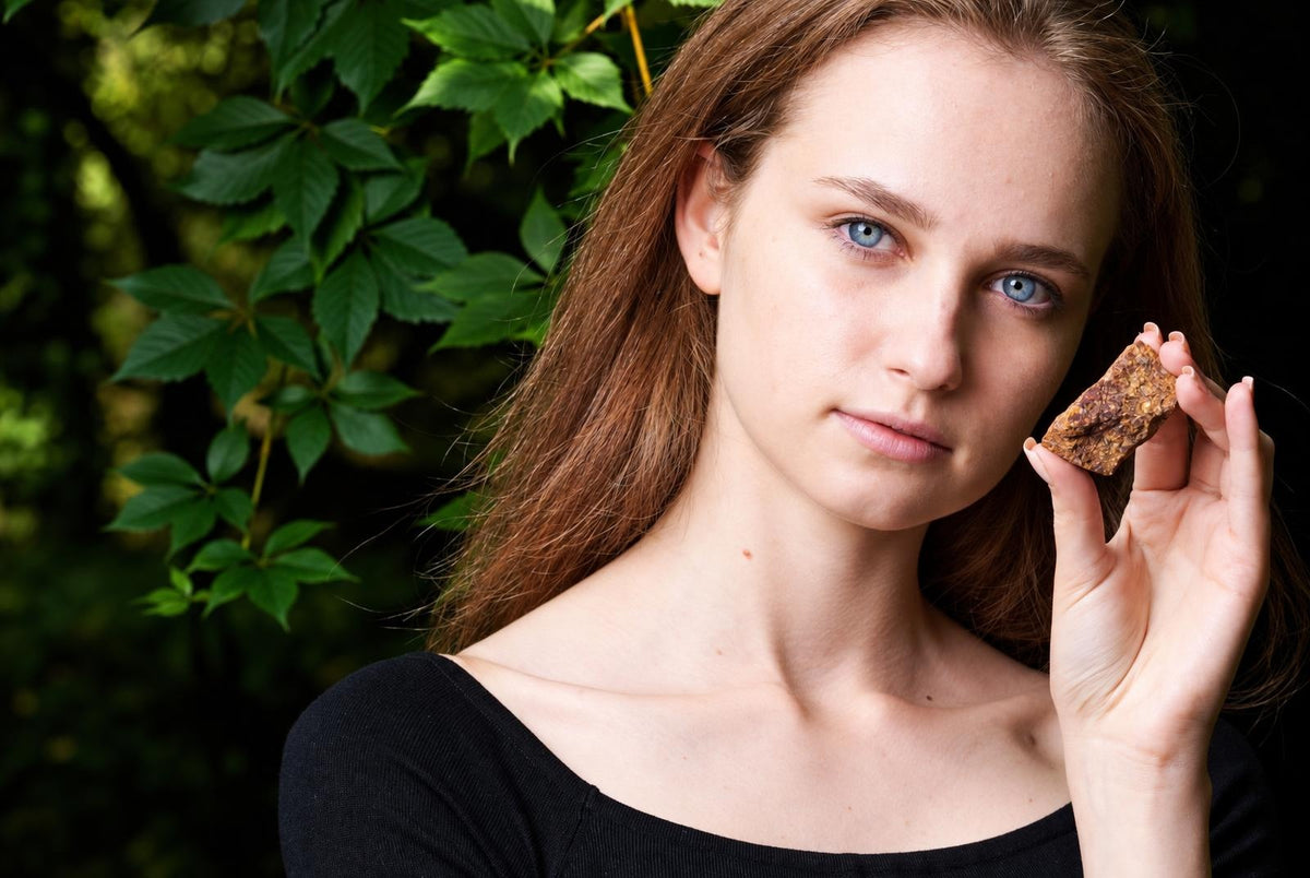 A woman with long brown hair and blue eyes holds a small, rough-textured brown stone near her face against green leaves.