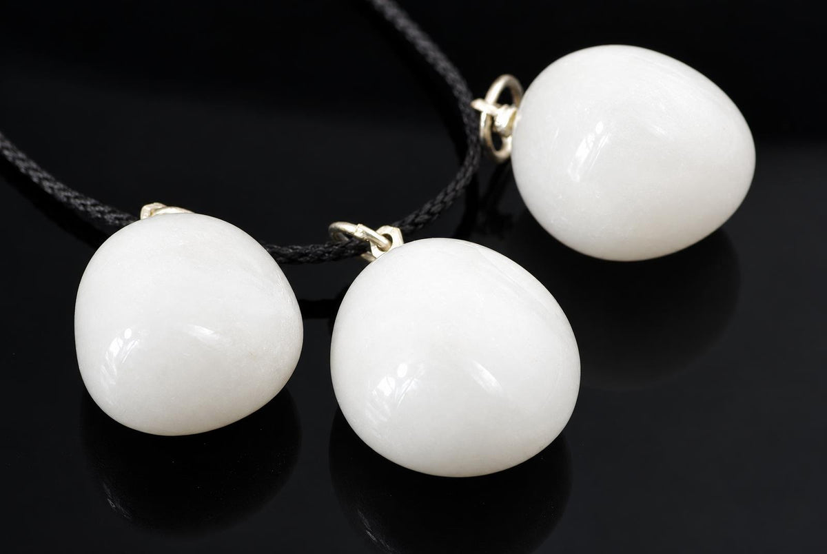 Smooth white snow quartz pebble pendants with metal loops and black cords are grouped together on a glossy dark surface.