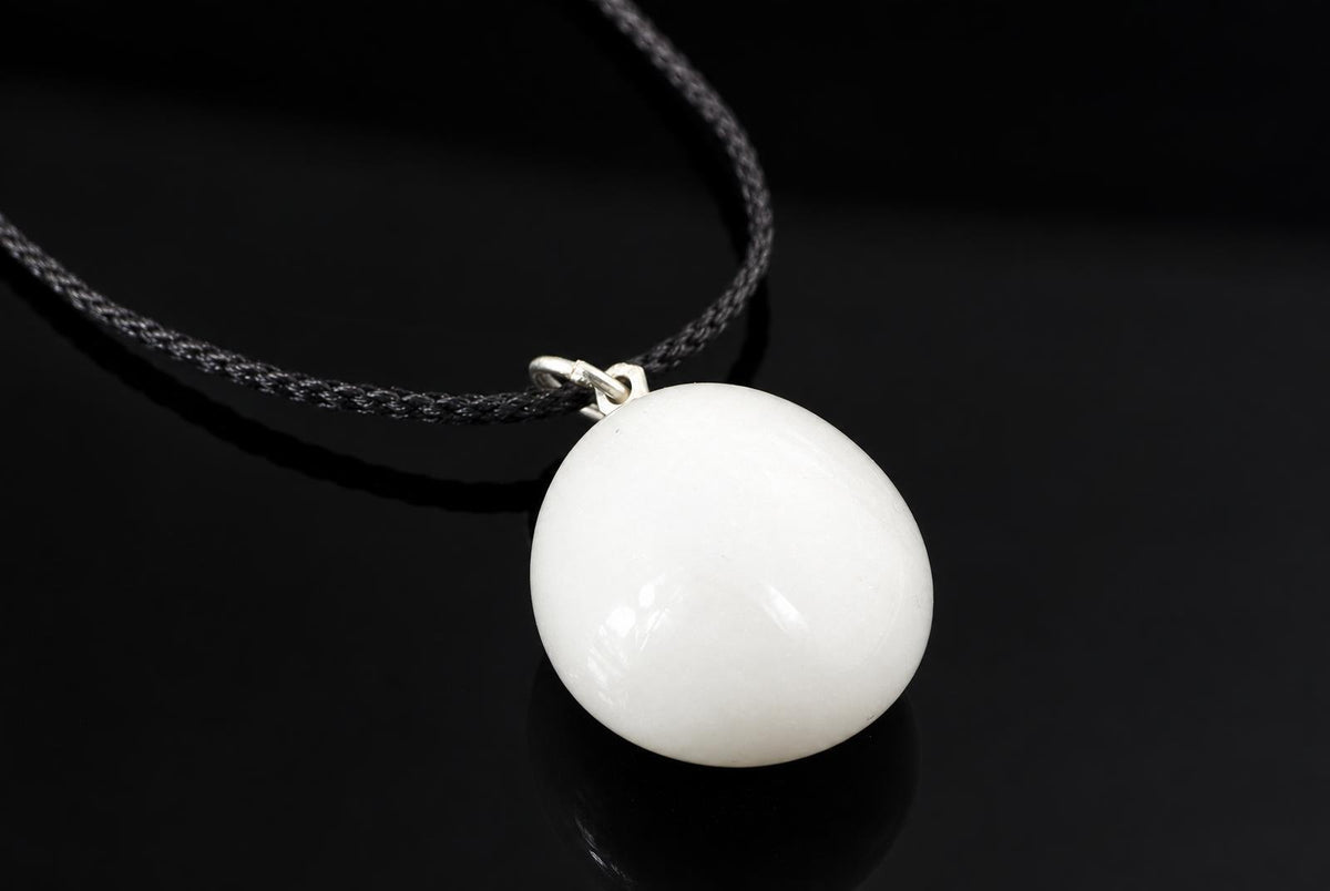 A smooth white snow quartz pebble pendant hangs from a black cord, with a soft reflection on the dark surface below.