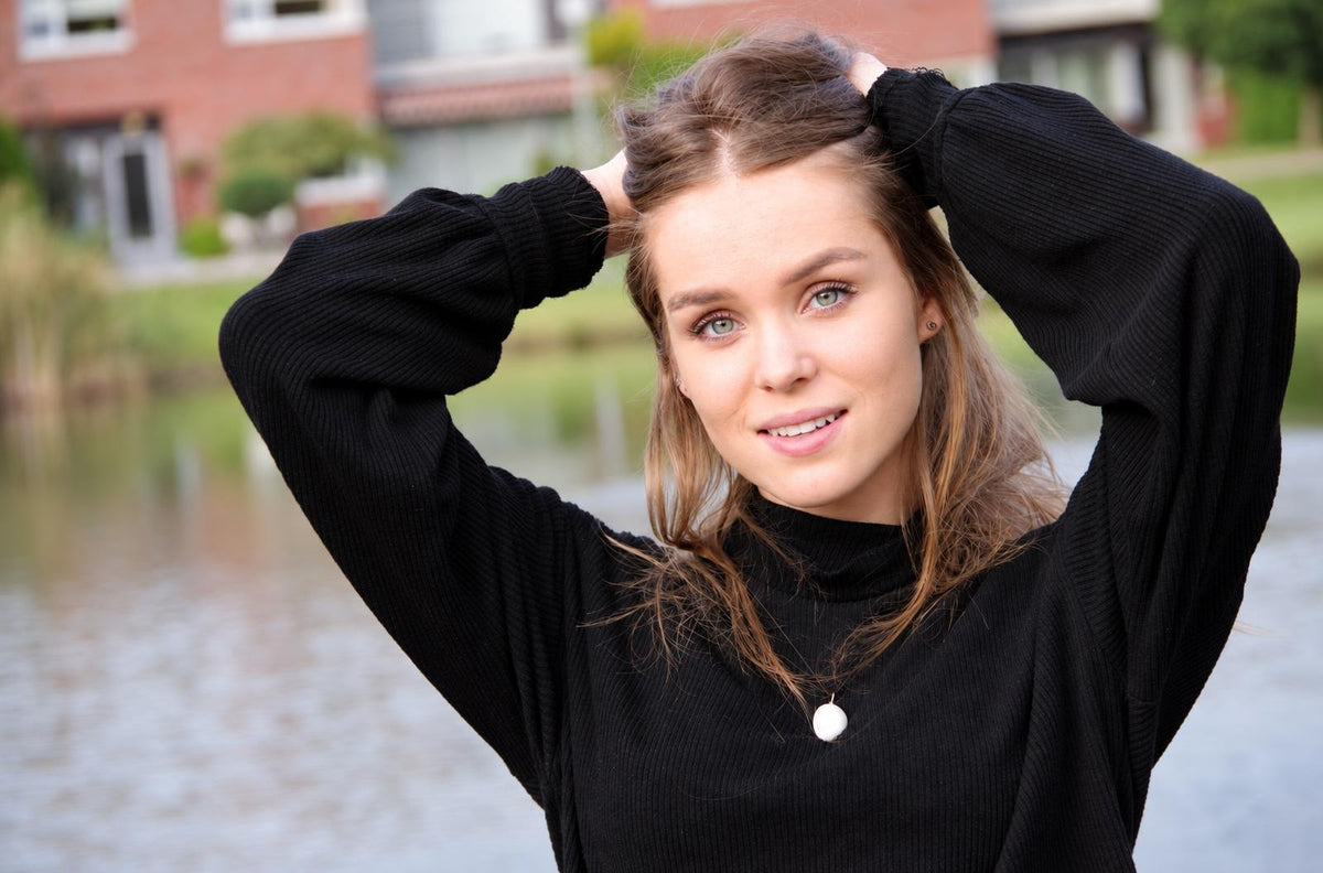 A woman in a black turtleneck wears a white round pendant necklace, hands in hair, outdoors near water.