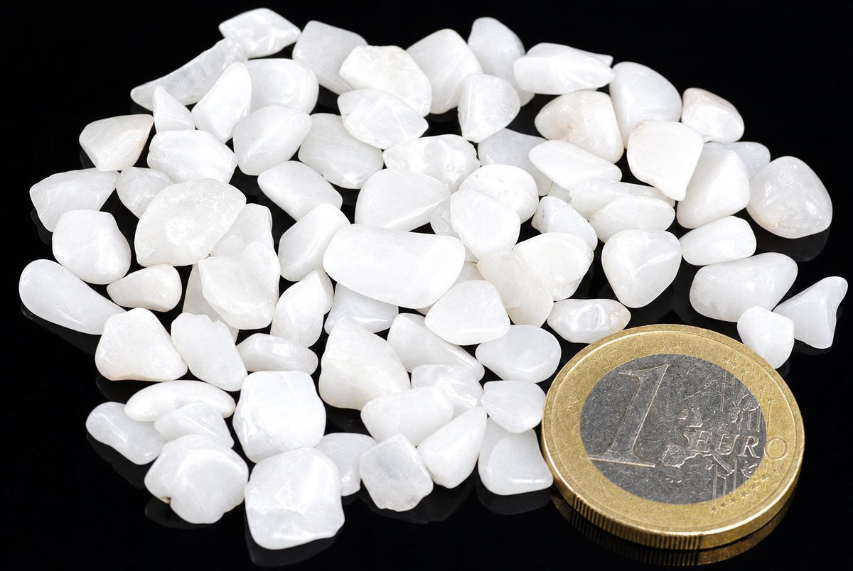 A pile of small, white, polished quartz stones on a dark surface next to a one euro coin.