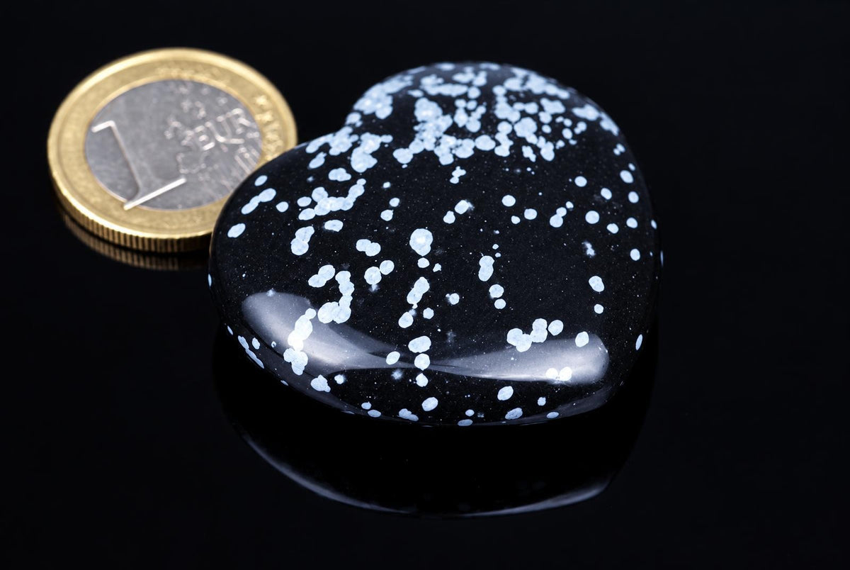 A polished black heart-shaped stone covered with pale speckles rests beside a coin on a reflective black surface.