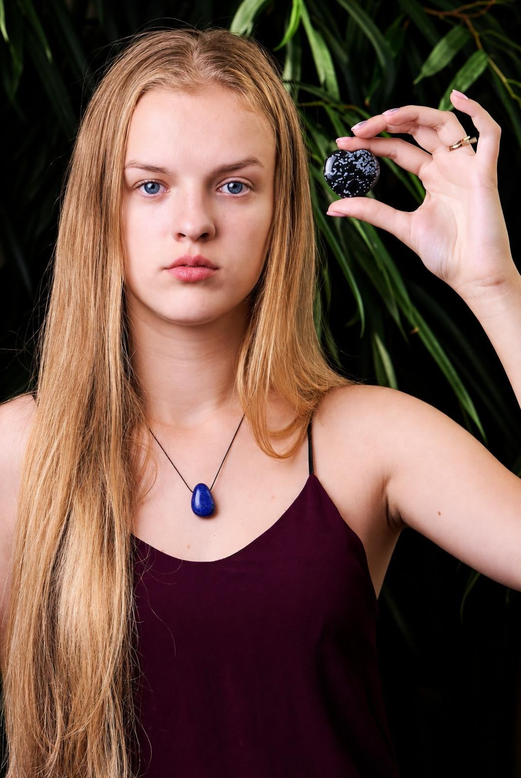 A woman holds a black, heart-shaped stone with white speckles against green leaves. She wears a blue pendant necklace and a dark top.