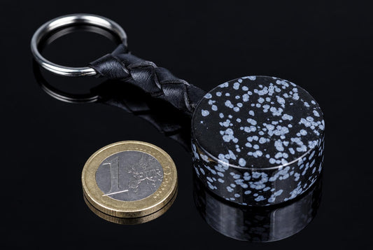 A black and white speckled cylindrical keychain with a braided leather strap and metal ring, next to a one-euro coin on a dark surface.
