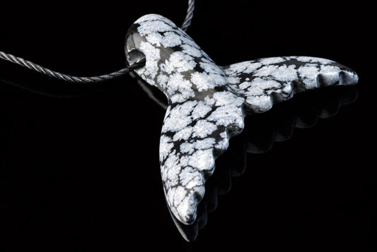 A carved pendant shaped like a dolphin tail, featuring white and black snowflake patterns on a dark surface, suspended from a cord against a black background.