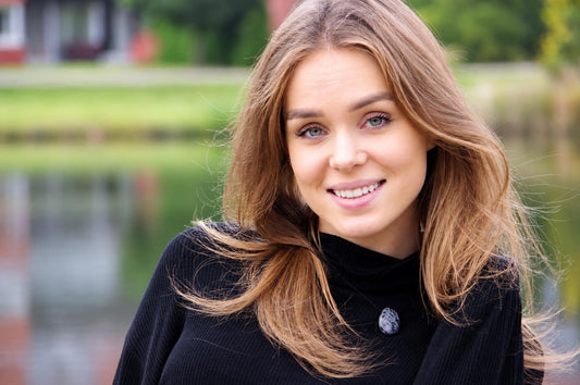 A woman with long brown hair wears a black turtleneck sweater featuring a round, dark grey pendant with white speckles.