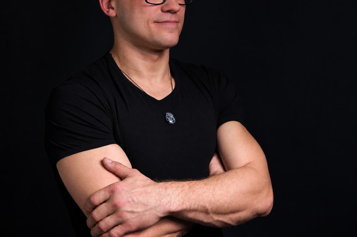 A man in a black t-shirt wears a dark pendant with a light pattern on a thin cord against a dark background.
