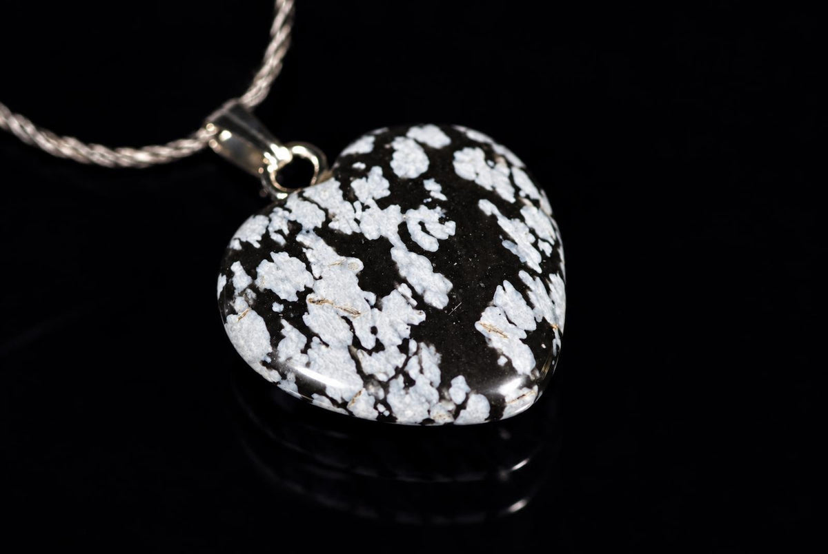 A heart-shaped pendant with a black and white speckled stone on a silver-toned chain, resting on a dark surface.