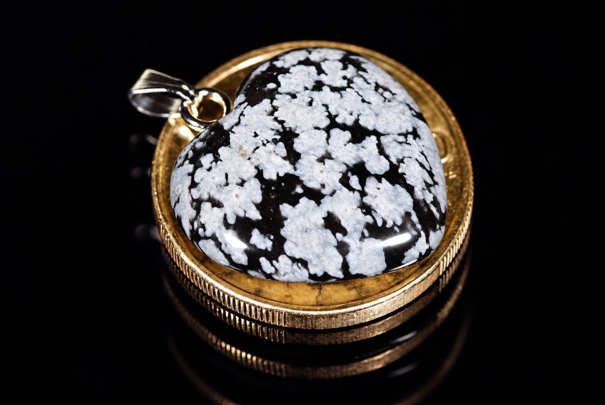 A black heart pendant with pale speckles and a metal loop rests on a coin against a black background.