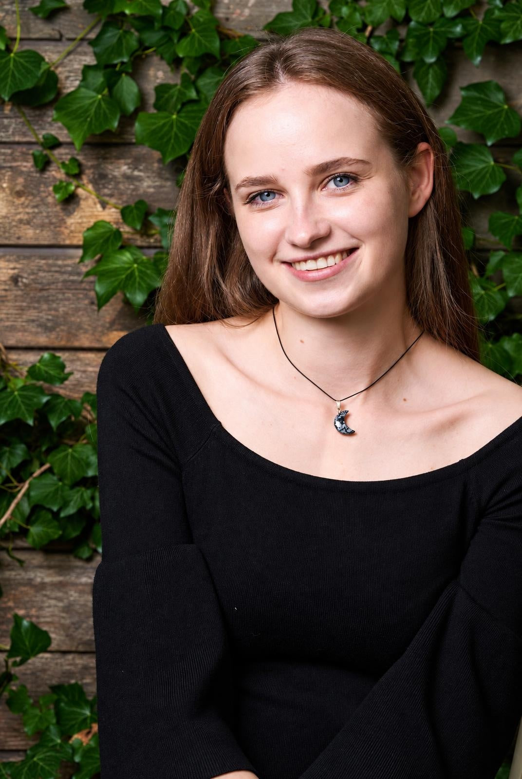 A woman wears a black top and a necklace with a dark crescent moon pendant on a thin cord, against a wooden wall with green ivy.