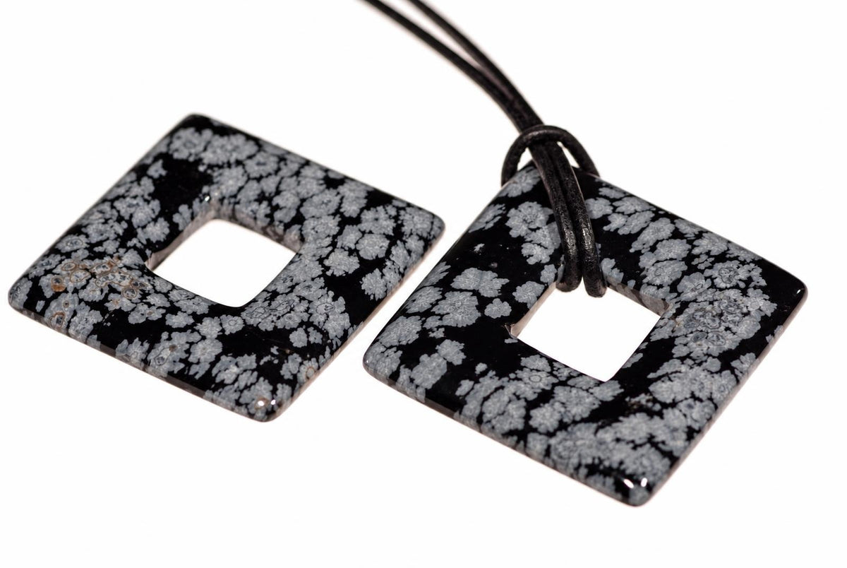 Two square, donut-shaped pendants with a black and white snowflake pattern lie on a pale surface; one has a dark cord attached.