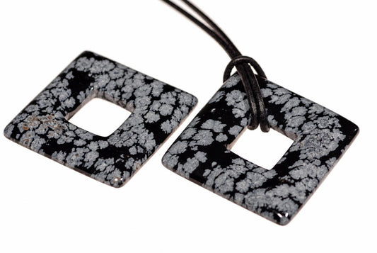 Two square, donut-shaped pendants with a black and white snowflake pattern lie on a pale surface; one has a dark cord attached.