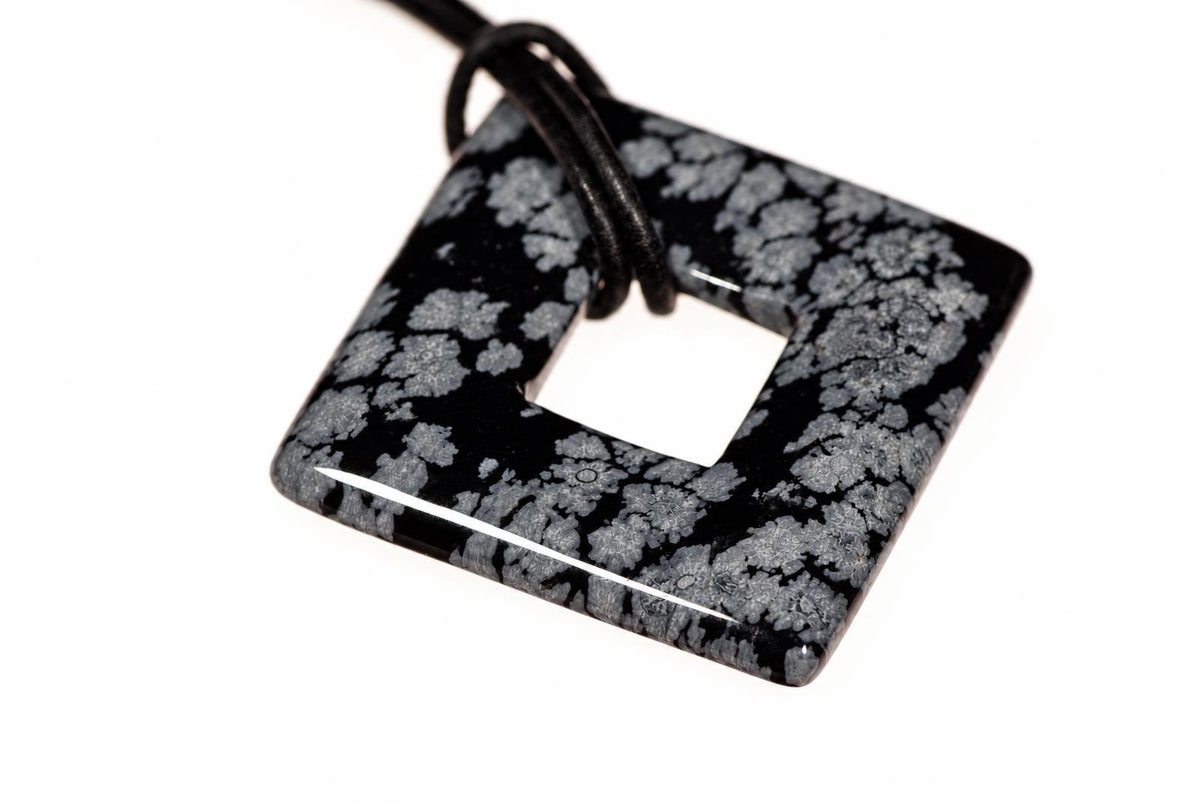 A square pendant with a central hole, featuring a black and grey snowflake pattern on a dark surface, attached to a black cord against a white background.