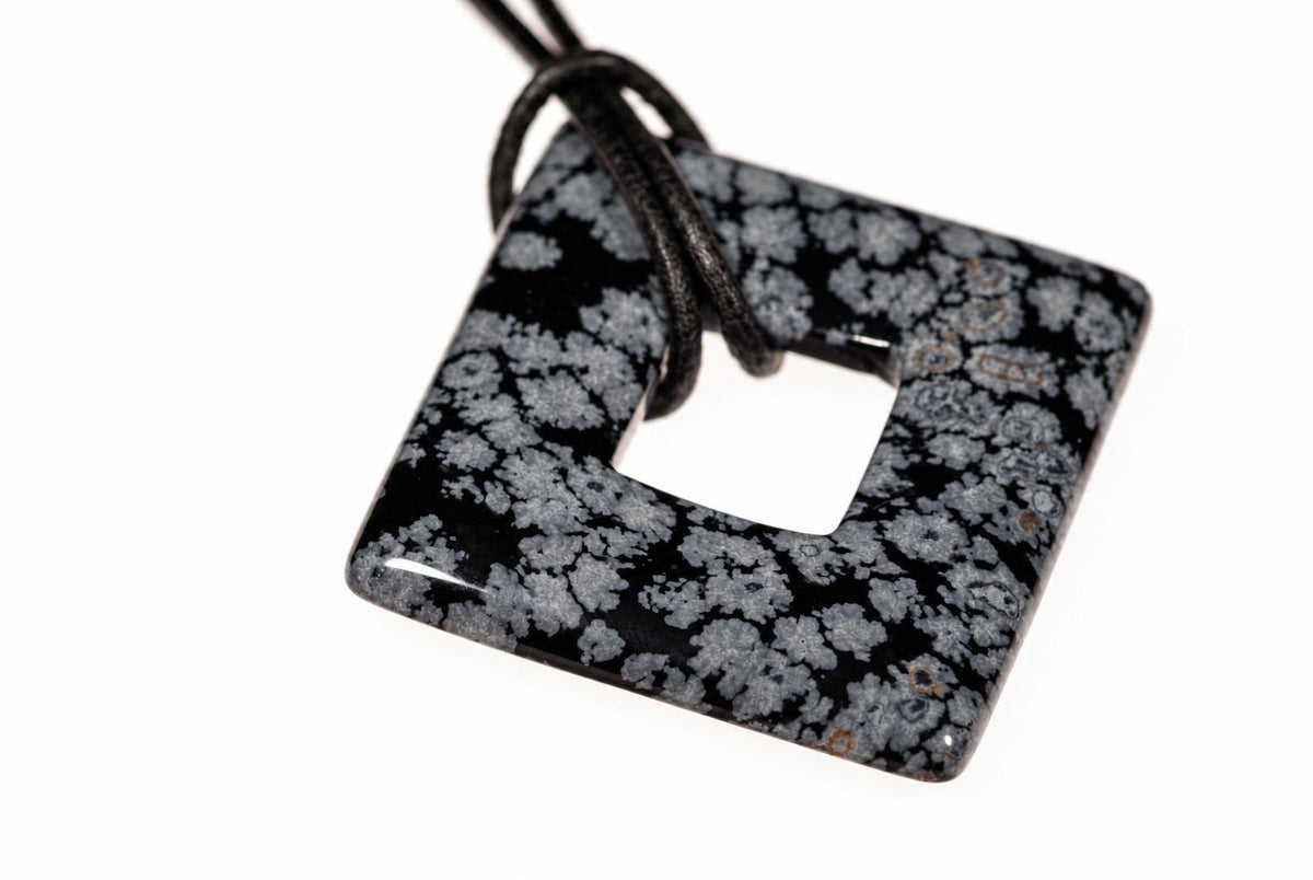 A square pendant with a central hole, featuring a black and white speckled pattern resembling snowflakes, attached to a dark cord against a white background.
