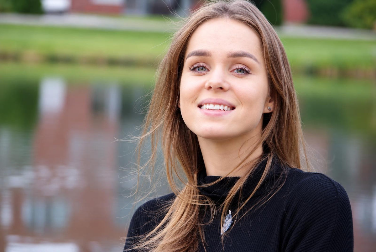 Portrait of a smiling woman with long brown hair wearing a black top and a silver-toned pendant necklace, outdoors near water.