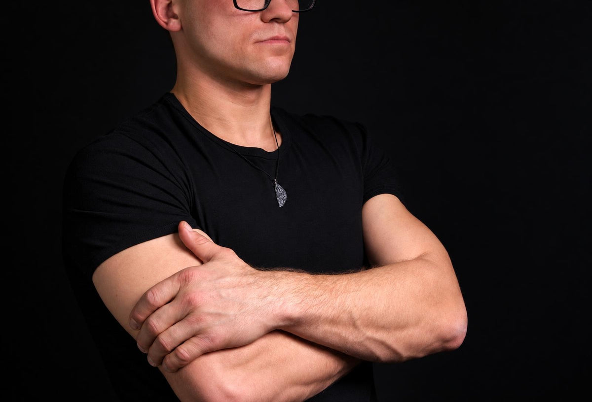 A man in a black t-shirt wears a dark pendant on a thin cord against a dark background.