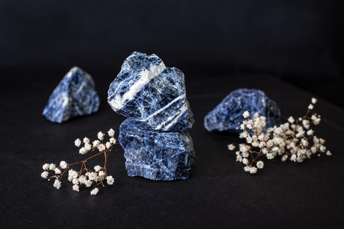 Three blue sodalite stones with white veins rest on a dark surface, accompanied by sprigs of dried baby's breath flowers.
