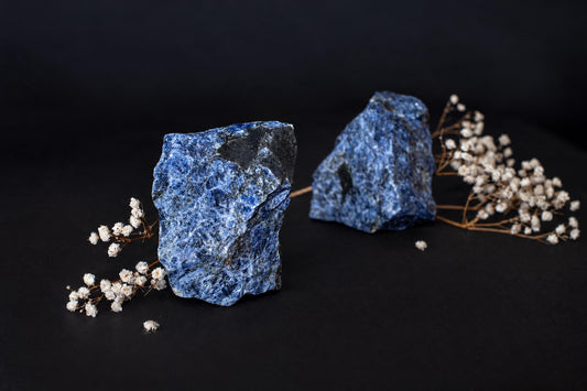 Two blue sodalite stones with black patches sit on a dark surface, flanked by sprigs of dried white flowers.