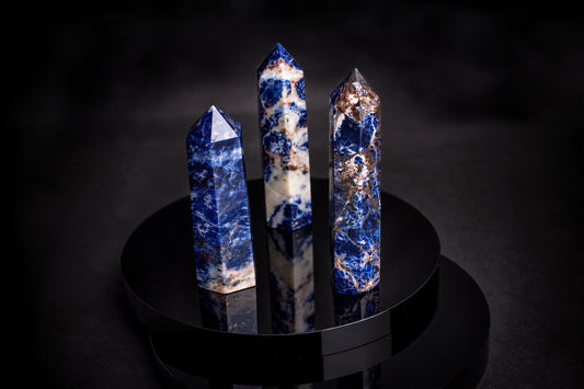 Three polished sodalite points with blue, white and brown patterns stand on a dark reflective surface.