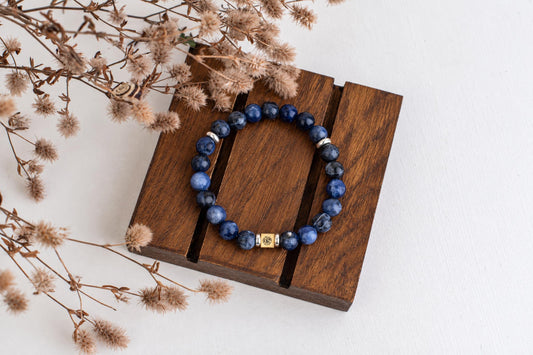 A blue beaded bracelet with silver and gold-toned accents rests on a dark wooden surface, surrounded by dried flowers.