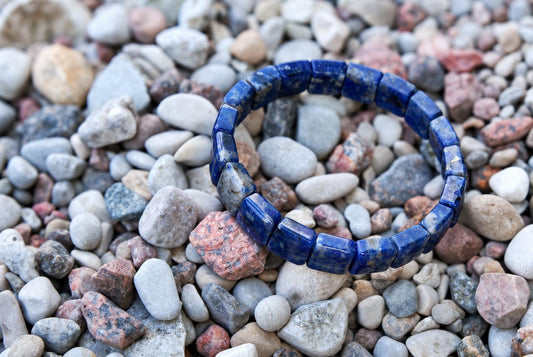 A dark blue sodalite bracelet with square beads rests on a bed of multicoloured pebbles.
