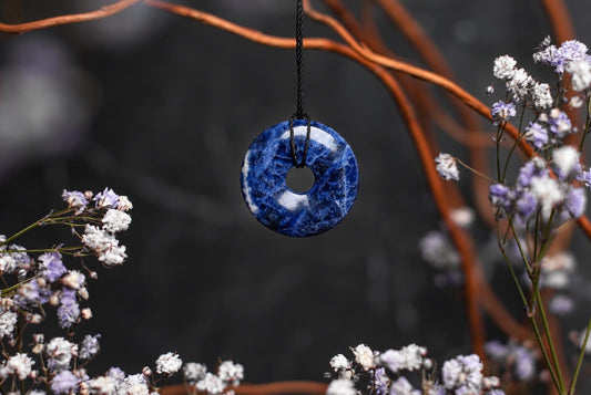 A blue sodalite donut-shaped pendant with white swirls hangs from a black cord against a dark background with flowers.