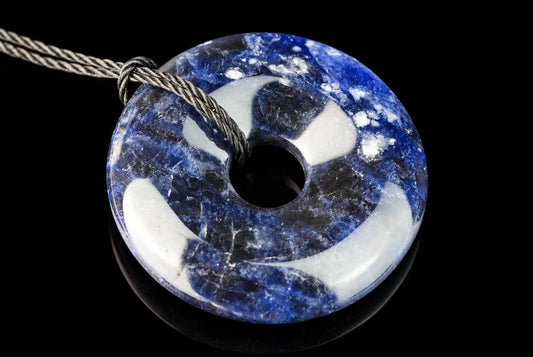 A polished sodalite pendant with blue and white swirls, shaped like a donut with a central hole, on a dark surface.