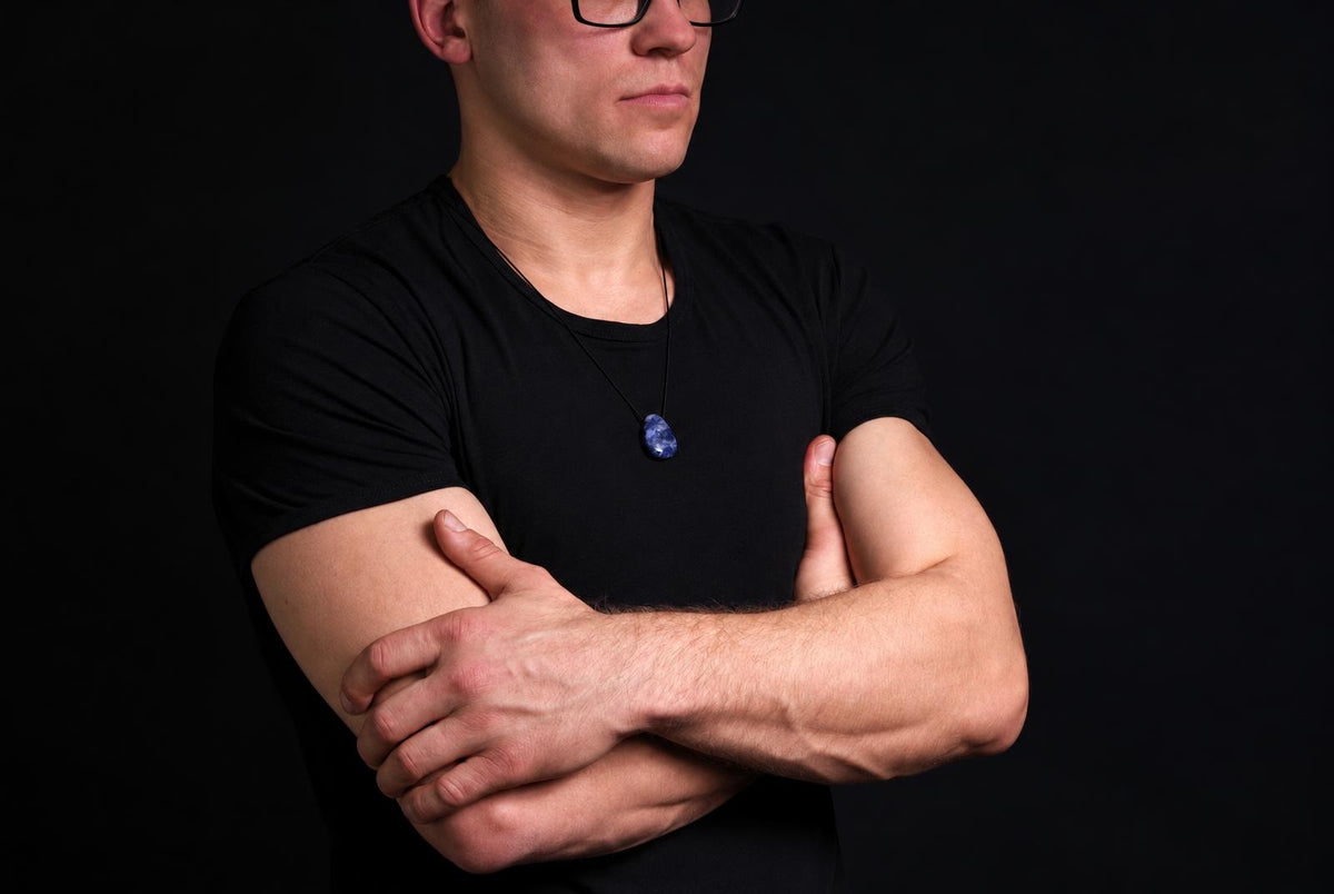 A man in a black shirt wears a necklace with an oval blue sodalite pendant on a dark surface.