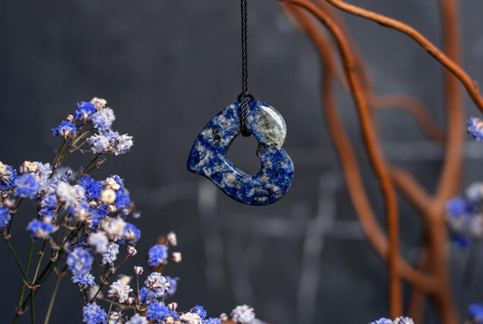 A polished sodalite pendant shaped like a heart hangs from a black cord against a dark background with blue flowers and brown branches.