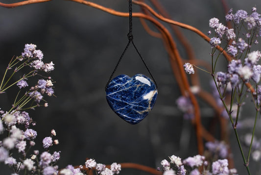 A heart-shaped sodalite pendant with white veins hangs from a black cord, surrounded by purple flowers and brown branches.