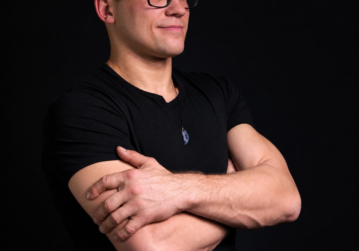 A man in a black t-shirt wears a dark blue and white patterned pendant on a thin cord against a dark background.